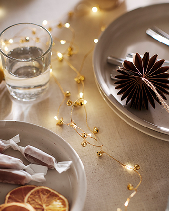 Mesa decorada para las fiestas con platos en tonos neutros, un vaso de cristal, caramelos envueltos, rodajas de naranja seca y una guirnalda de luces cálidas. Un adorno de papel plegado aporta un toque festivo y elegante.