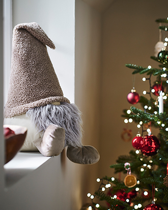 Figura decorativa de gnomo de tela en tonos beige y gris, situada junto a un árbol de Navidad adornado con bolas rojas y luces cálidas.