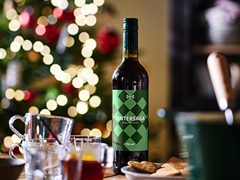 Botella de bebida VINTERSAGA sobre una mesa preparada para compartir, con vasos y aperitivos, delante de un árbol de Navidad iluminado.