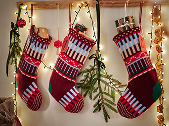 Calcetines navideños de punto en tonos rojos y blancos colgados en una pared decorada con ramas de abeto, luces y adornos festivos.