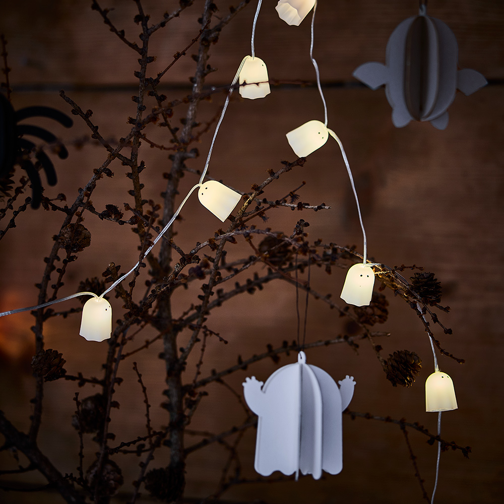 Una decoración colgante KUSTFYR con forma de fantasma blanco, suspendida de una rama, junto con una guirnalda de luces LED KUSTFYR con motivos de fantasmas.