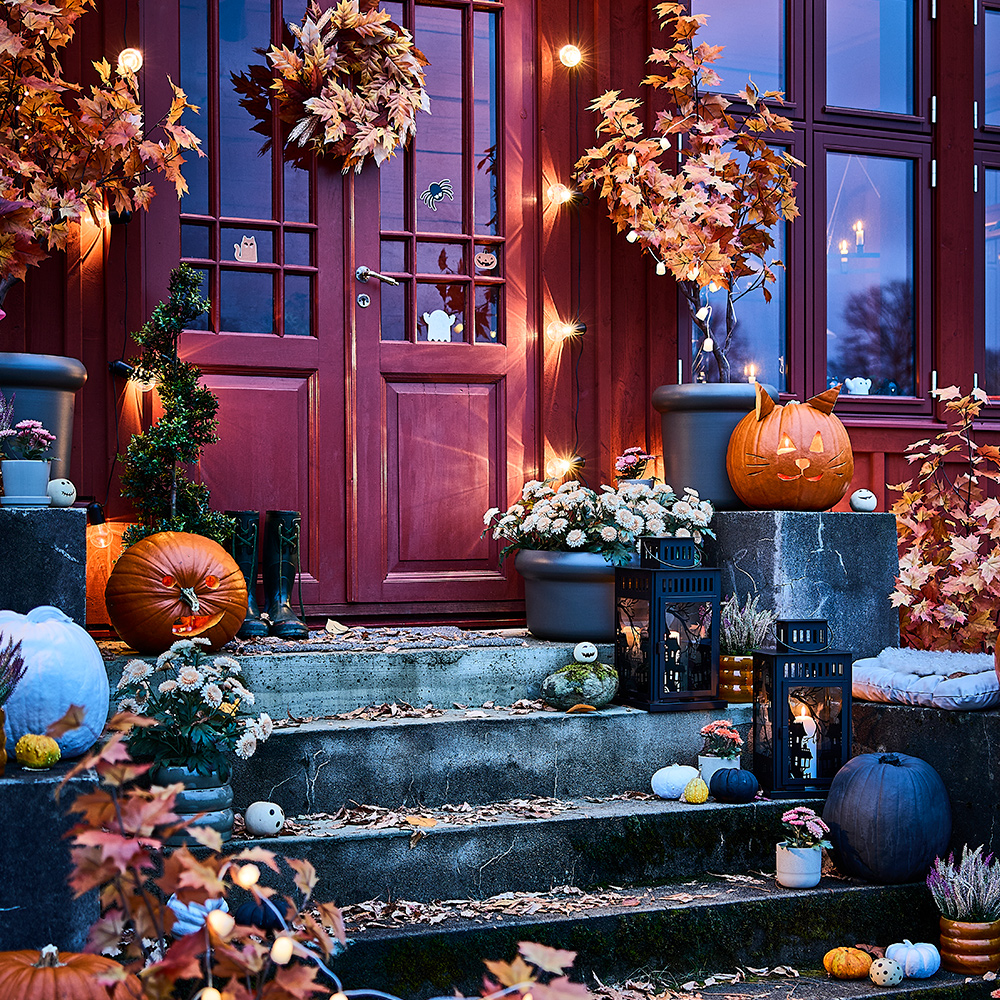 Una entrada de hogar iluminada y decorada para Halloween, con escaleras adornadas con calabazas y lámparas colgantes LED y faroles KUSTFYR.