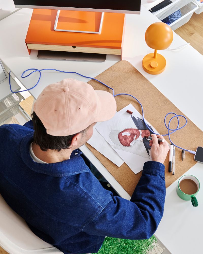 Person drawing an illustration at a desk with art supplies, an orange lamp and a cup of coffee.