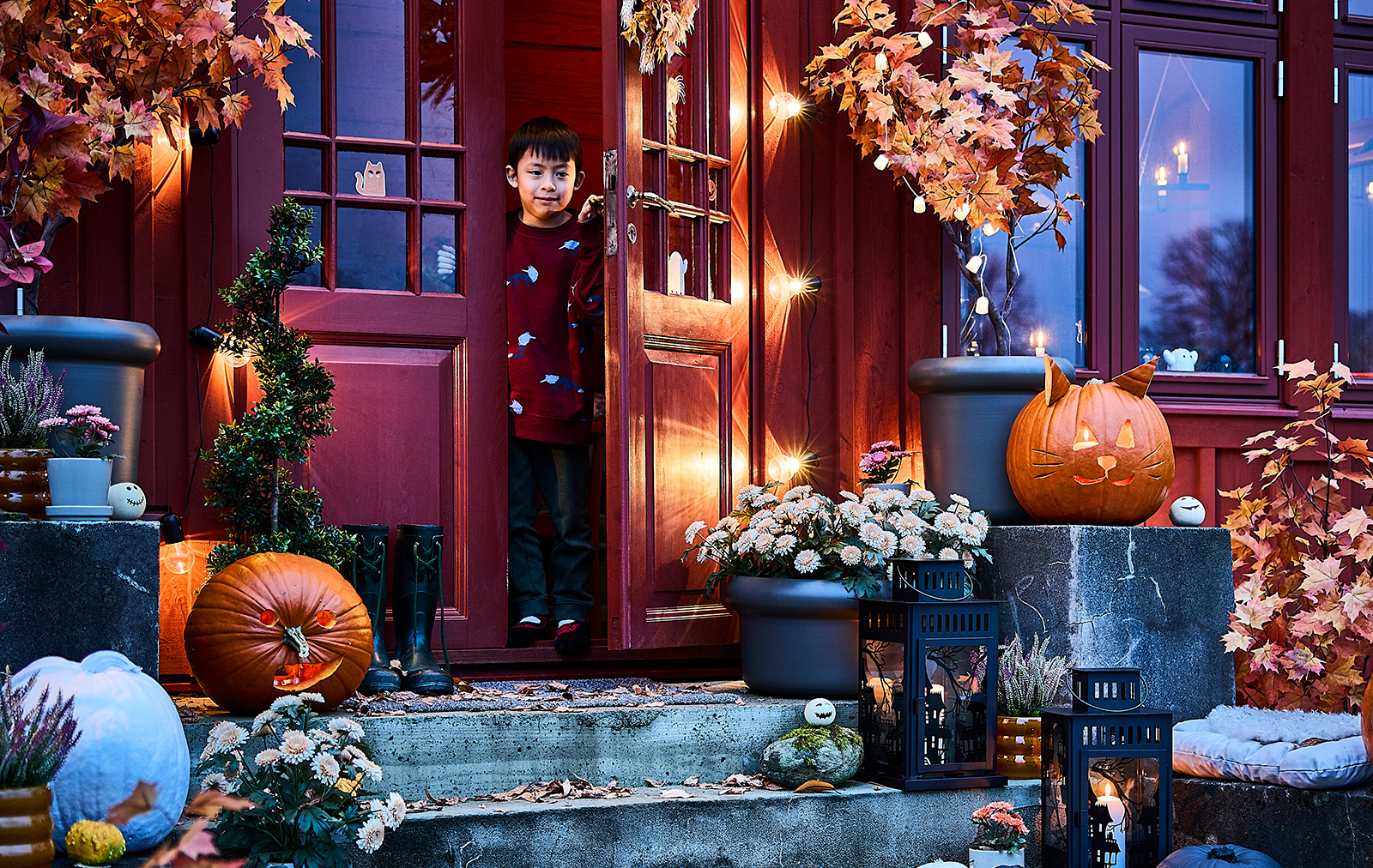 Un niño en la puerta abierta de una entrada iluminada y decorada para Halloween, utilizando lámparas colgantes LED y faroles KUSTFYR.
