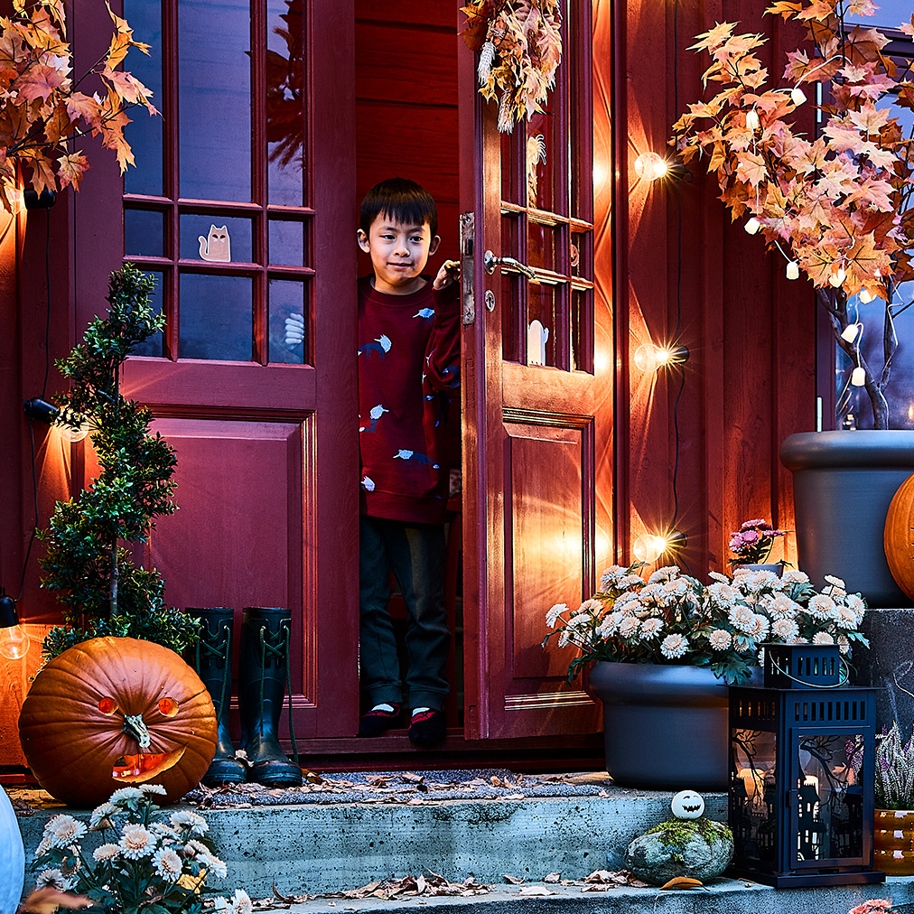 Un niño en la puerta abierta de una entrada iluminada y decorada para Halloween, utilizando lámparas colgantes LED y faroles KUSTFYR.