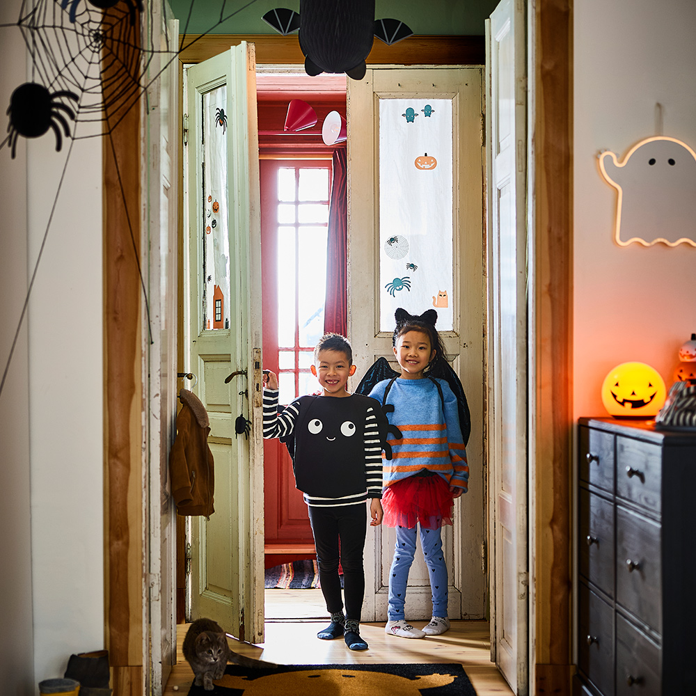 Dos niños están de pie en la entrada de un pasillo decorado con temática de Halloween, que cuenta con un felpudo KUSTFYR y decoraciones colgantes.
