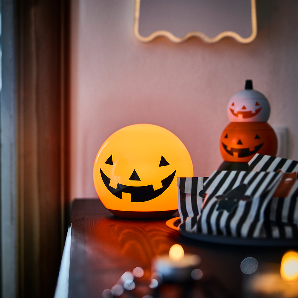 Un aparador exhibe una lámpara de mesa LED KUSTFYR con forma de calabaza iluminada y un conjunto apilado de tres latas con forma de calabaza.