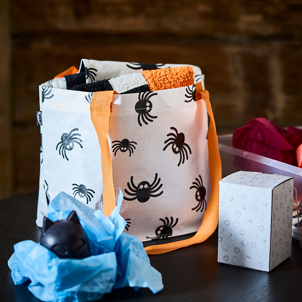Una bolsa de transporte KUSTFYR blanca con estampado de arañas sobre una mesa, junto a una vela KUSTFYR con forma de gato negro envuelta en papel de seda.
