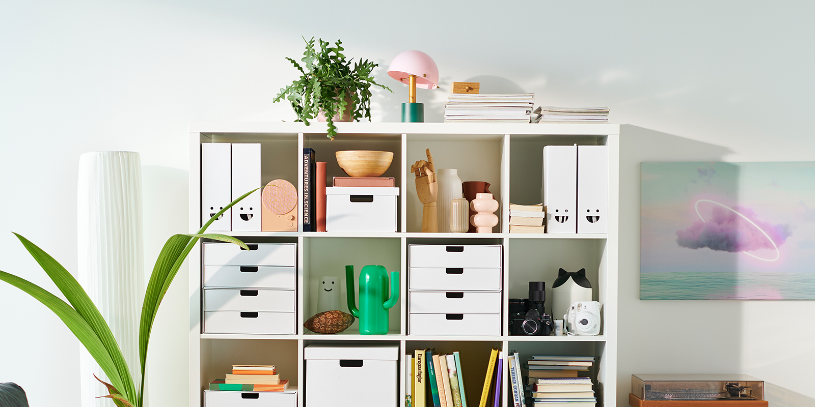 White IKEA KALLAX shelf unit organized with magazine files, storage boxes, books, plants, and decorative accessories in a bright room.