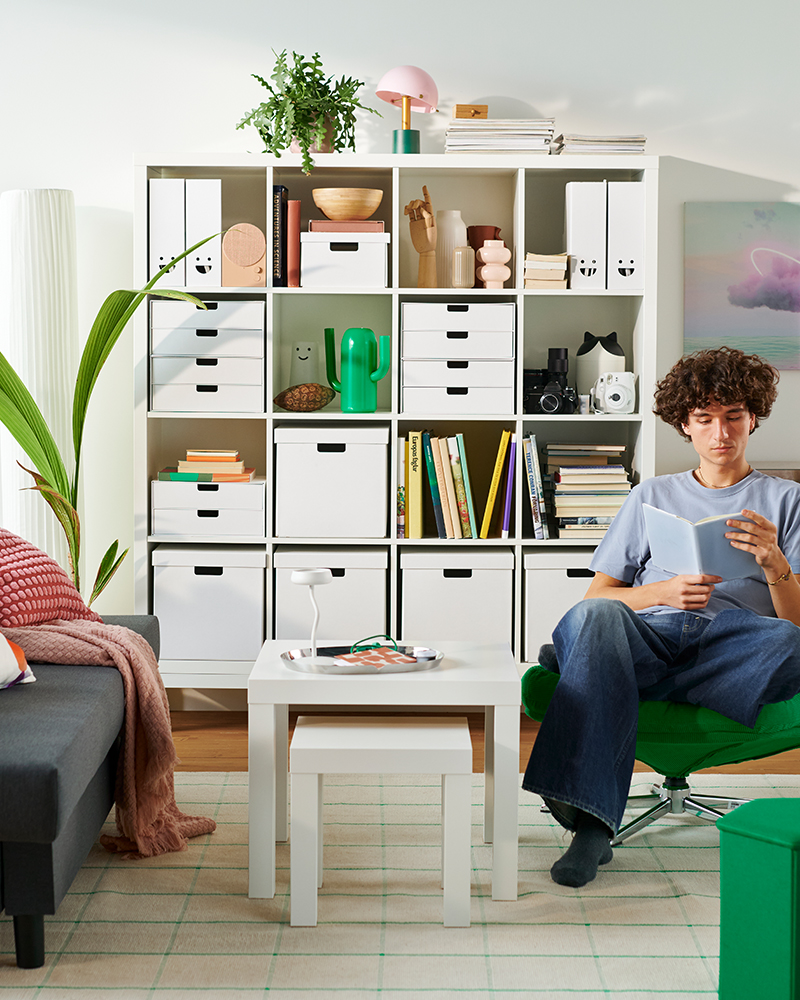 A living room with a KALLAX shelving unit filled with books and decorative items. A person is sitting in an armchair reading.