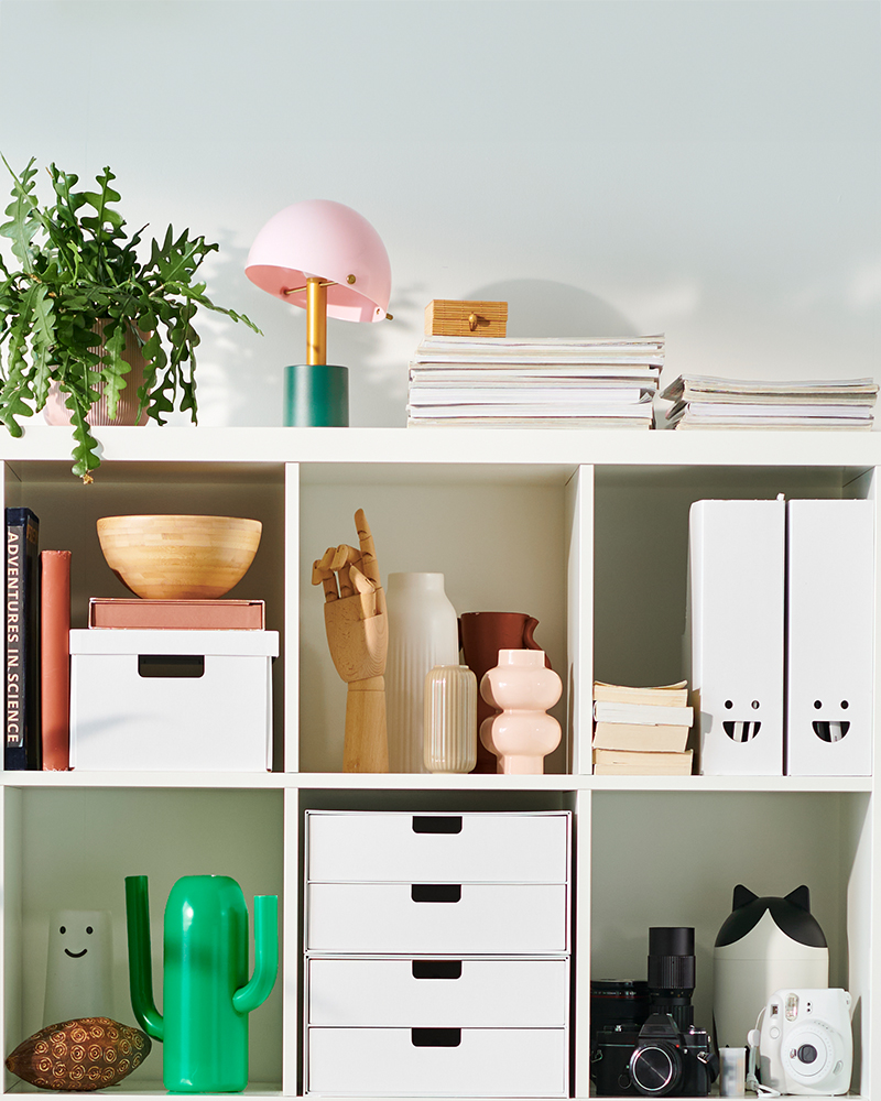 White IKEA KALLAX shelf unit organized with magazine files, storage boxes, books, plants, and decorative accessories in a bright room.