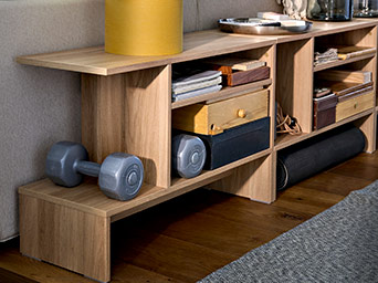 Wooden modular shelving unit storing dumbbells, a rolled yoga mat, and small storage boxes in a cozy living room space.