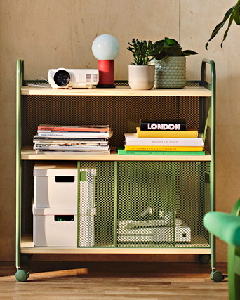 Green wheeled utility cart with mesh shelves, neatly organized with books, magazines, small storage boxes, and potted houseplants.