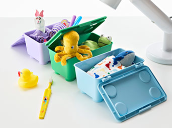 Colorful plastic storage boxes with lids holding soft plush toys and children's items, placed on a white desk next to a rubber duck.