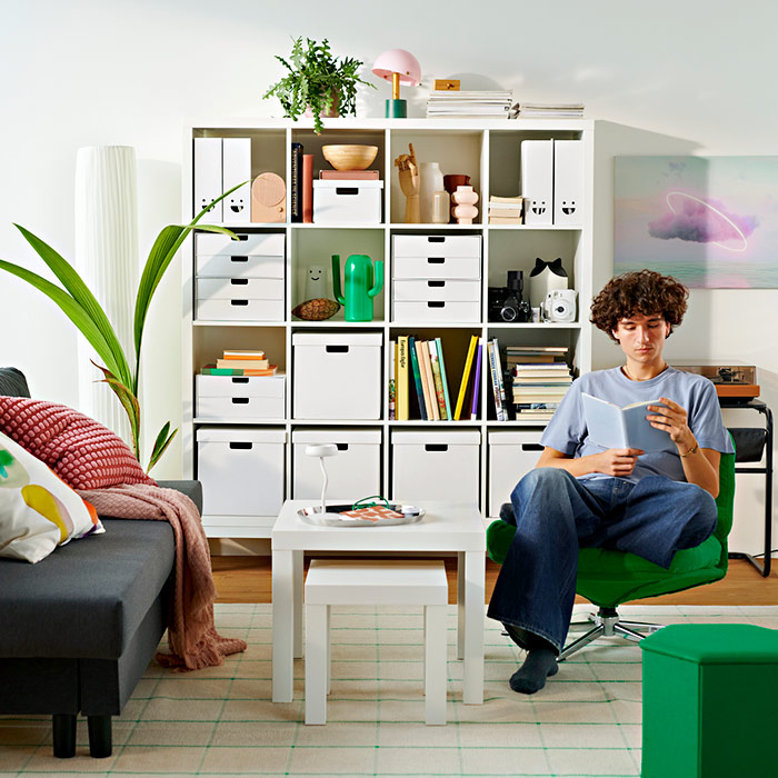 Young man reading a book in a green armchair in a bright living room with a large white grid shelving unit full of books and boxes.