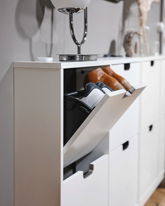 White shoe cabinet with a tilt-out compartment open, revealing shoes stored neatly inside near a table lamp.