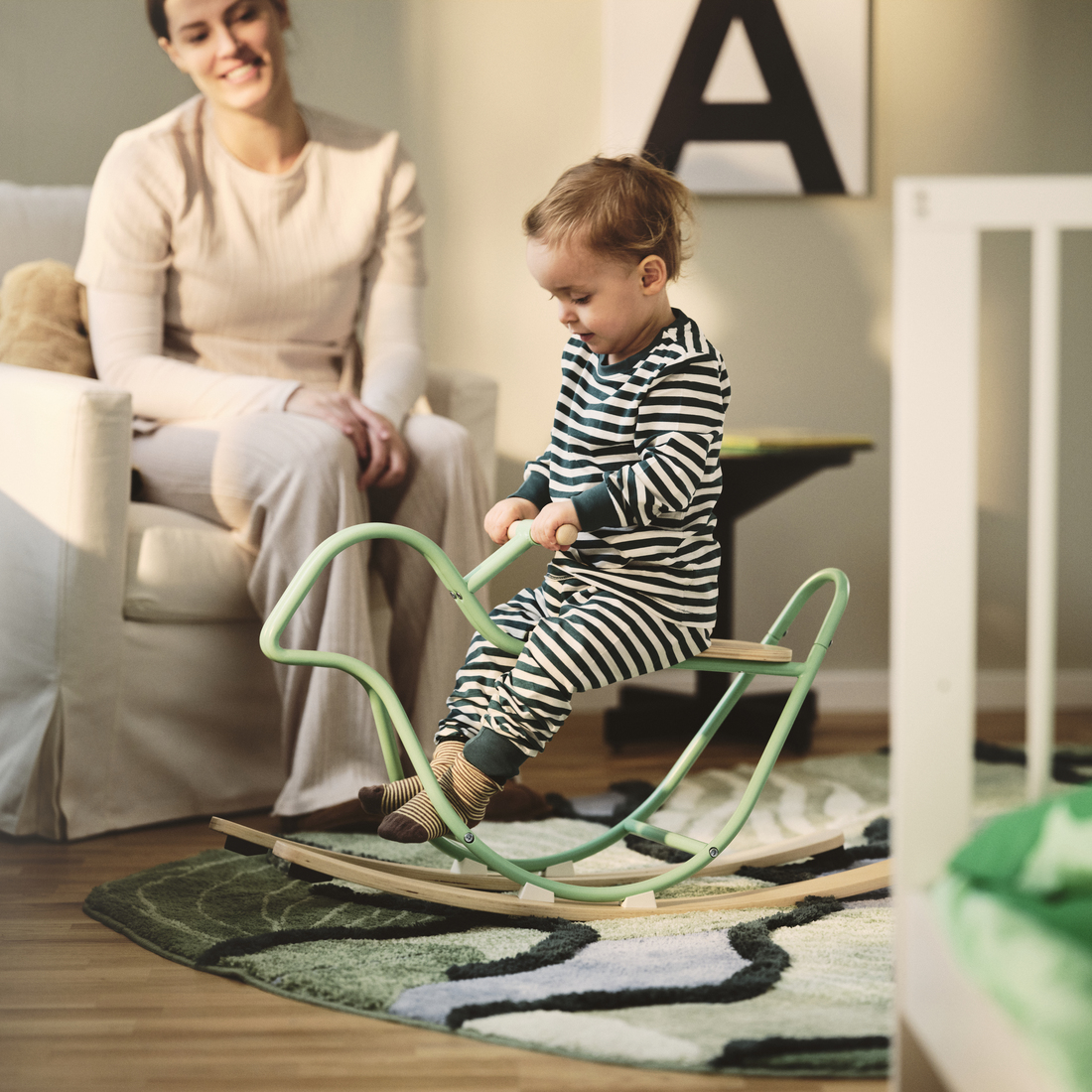 Toddler in striped pajamas riding a mint-green rocking moose over a green patterned rug, watched by an adult in a beige armchair.