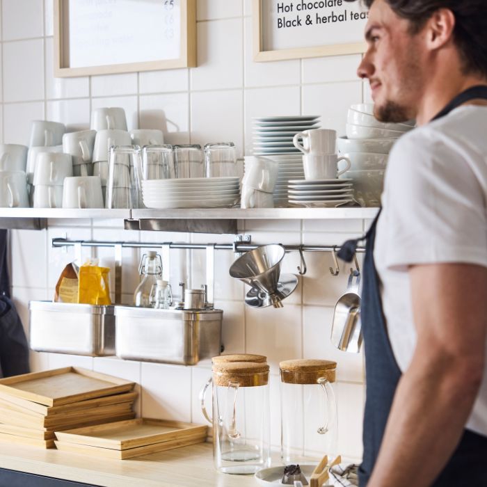 Close-up of café counter with stacked cups, glassware, and barista in apron; shelves hold jars, trays, and hanging tools.