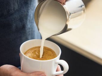 Milk poured from small pitcher into coffee cup, creating foam, with hands holding cup and pitcher in café setting.