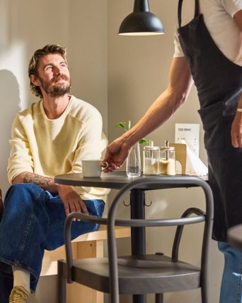 Barista serves coffee at small café table as seated customer looks up; cups, sugar jars, and warm light create cozy setting.