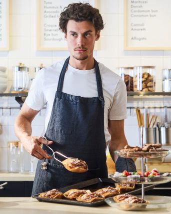 Barista in apron plating pastries behind café counter, holding tongs; trays, jars, and menu boards in bright kitchen setting.