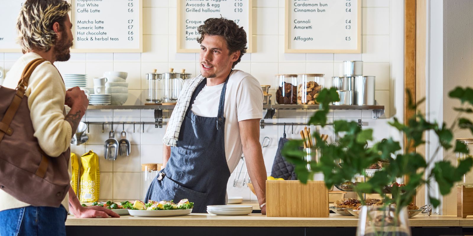 Barista behind café counter chats with customer; menu boards, cups, and pastries visible in bright, modern interior.