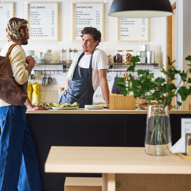 Barista behind café counter chats with customer; menu boards, cups, and pastries visible in bright, modern interior.
