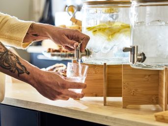 Hands fill a glass from a water dispenser with lemon slices and ice on a wooden stand, set on a café counter.