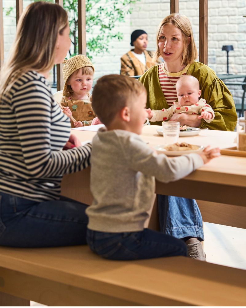 Adults and children sit at café table by window, with drinks and snacks; one holds infant while others talk in bright space.