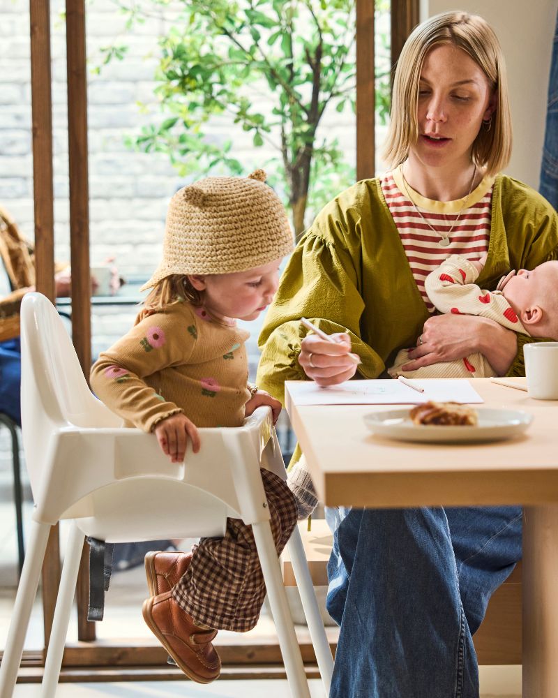 Adult seated at café table holding infant while assisting child in high chair; plate, cup, and window view in bright space.