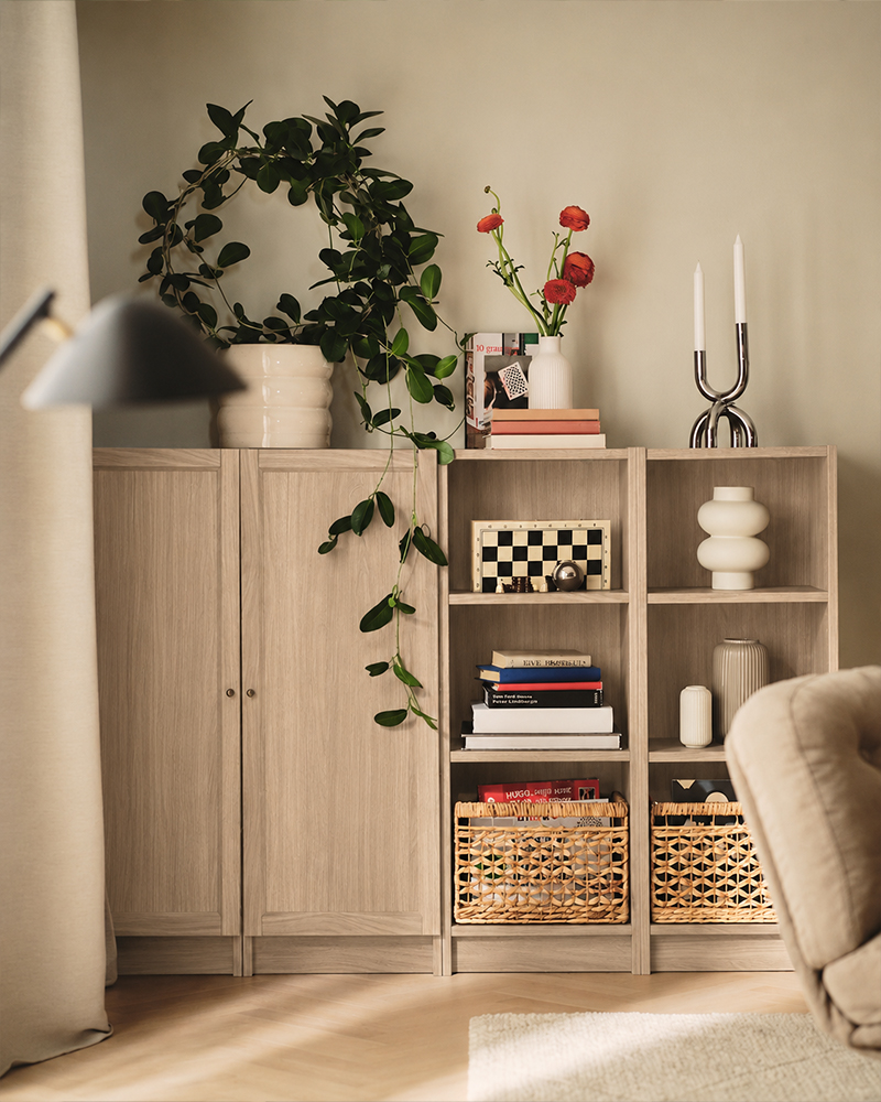 Modern living room divided by tall dark wood shelving, with a yellow sofa, white side table, and workspace in the background.