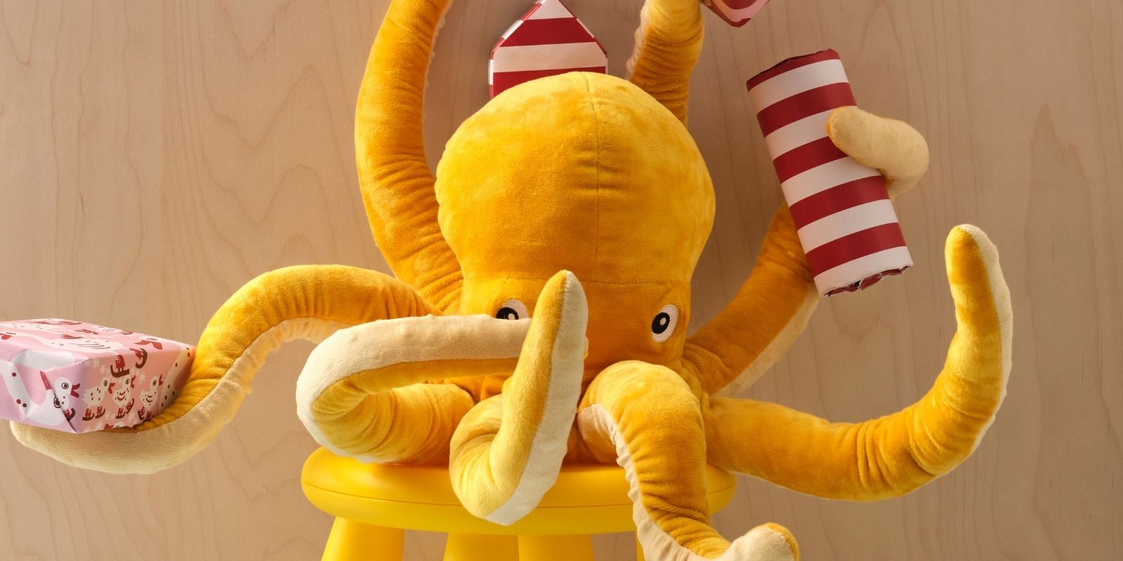 Un pulpo de peluche amarillo BLÅVINGAD está sentado en un taburete MAMMUT, sosteniendo varios regalos envueltos en cinco de sus ocho brazos.