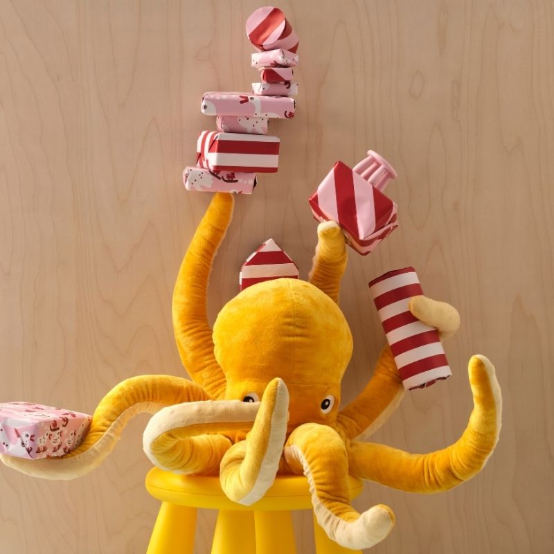 Un pulpo de peluche amarillo BLÅVINGAD está sentado en un taburete MAMMUT, sosteniendo varios regalos envueltos en cinco de sus ocho brazos.