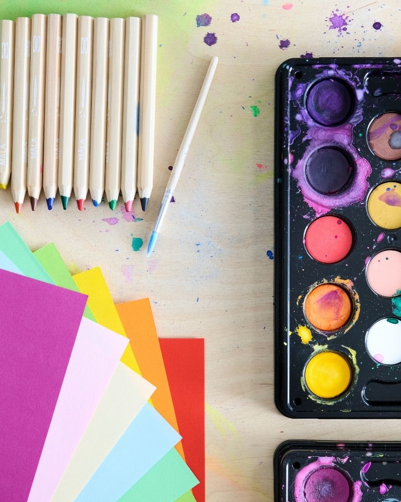 Papel de colores, un juego de lápices MÅLA y una caja de acuarelas, todos colocados sobre una mesa de madera. Foto tomada desde arriba.