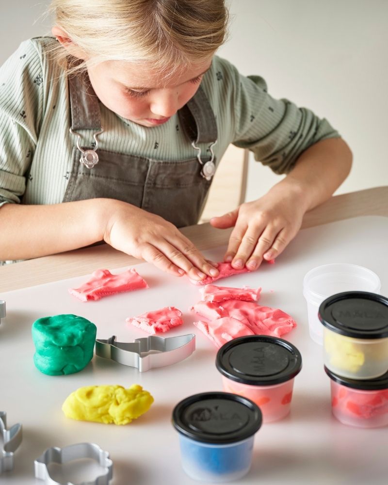 Una niña pequeña está sentada a la mesa, esculpiendo figuritas con plastilina de colores MÅLA.