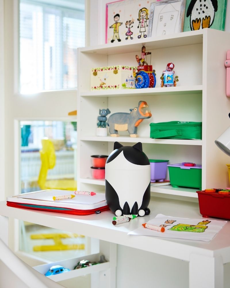 En una habitación infantil luminosa, un organizador con tapa GREJSIMOJS en forma de gato está sobre un escritorio junto a cajas de colores y otros objetos.