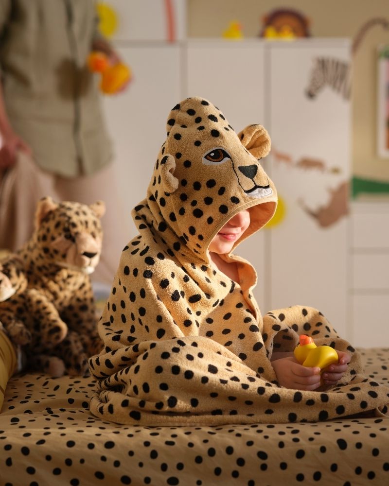 Un niño con una toalla con capucha SANDLÖPARE con estampado de guepardo está sentado en una cama rodeado de guepardos de peluche y un patito de goma.