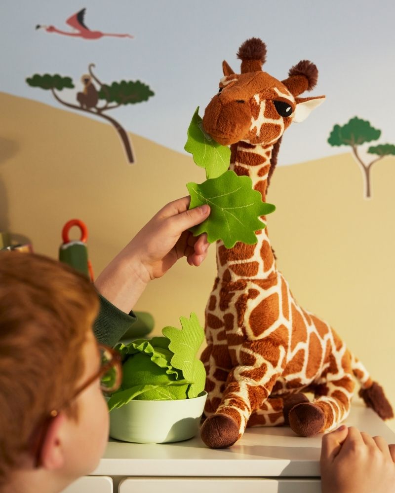 Un niño sostiene una hoja verde para alimentar a una jirafa de peluche SANDLÖPARE.