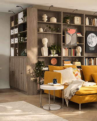 Modern living room divided by tall dark wood shelving, with a yellow sofa, white side table, and workspace in the background.