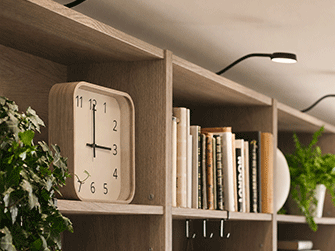 Square light wood clock on a dark walnut shelf next to books, green leafy plants, and top spotlighting.