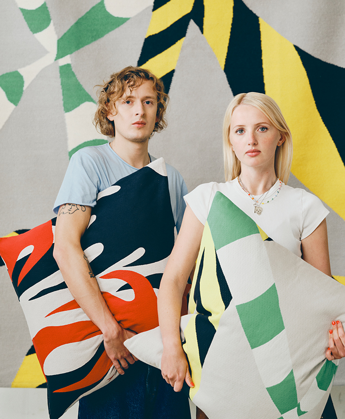 Two people pose holding decorative cushions with abstract patterns in green, black, white, yellow, and red. In the background, a rug or textile featuring the same geometric design creates a coordinated home furnishing display.