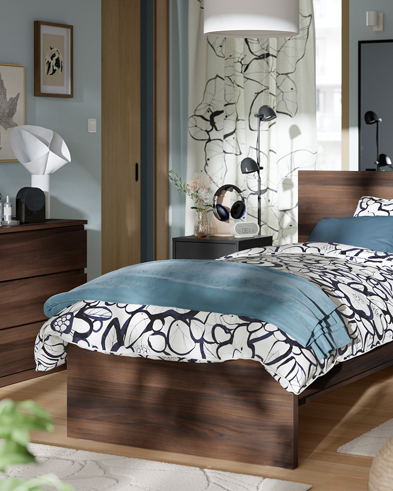 Cama individual STORKLINTA efecto madera oscura con cabecero y ropa de cama azul y estampada en un dormitorio moderno.