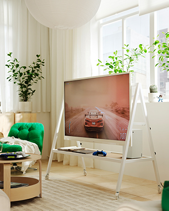 A white YXSTABY gaming TV stand on castors is placed in front of a window in a light living room. The TV shows a car game.