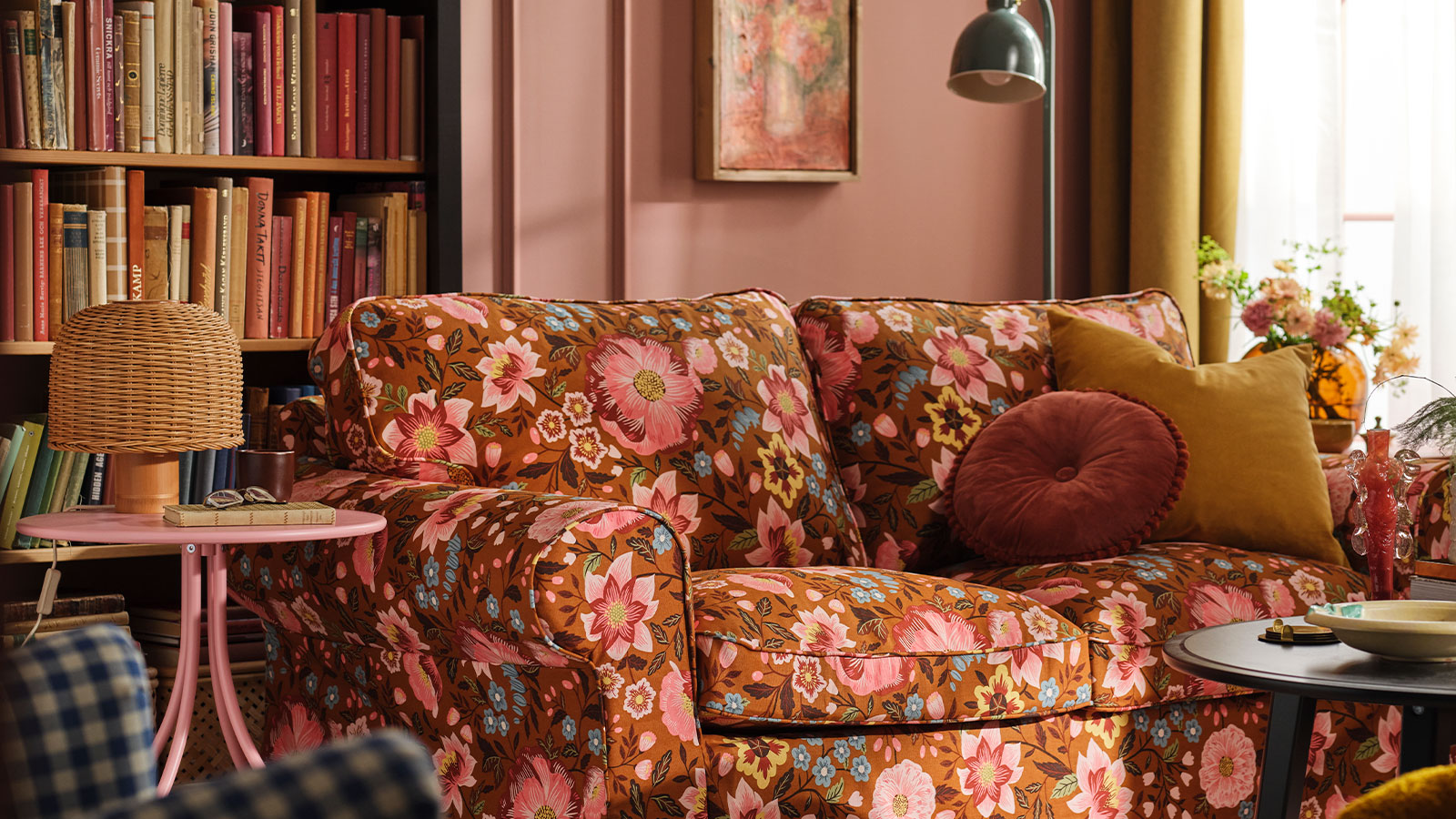 Un salón rosa con un sofá EKTORP cubierto con una funda Månggbyn marrón y multicolor, situado frente a una estantería.