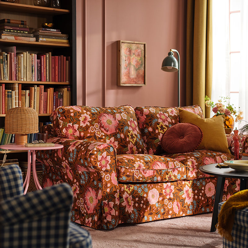 Un salón rosa con un sofá EKTORP cubierto con una funda Månggbyn marrón y multicolor, situado frente a una estantería.
