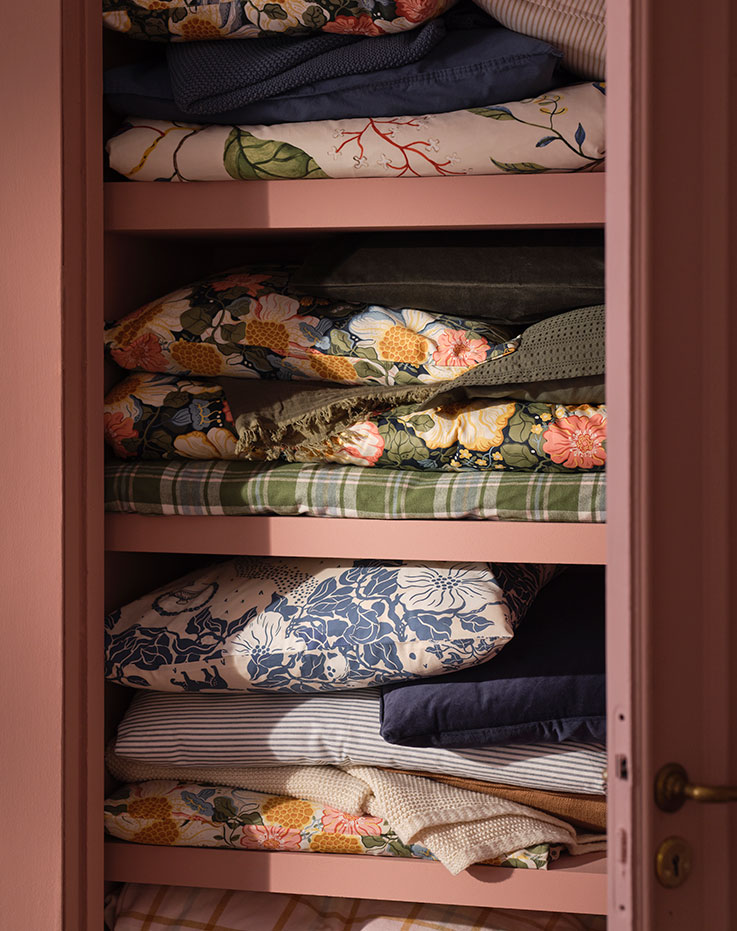 Un armario de color rosa con ropa de cama doblada, entre la que se incluyen fundas nórdicas y fundas de almohada de las colecciones PJÄXE y ARENBOK con diferentes estampados florales.