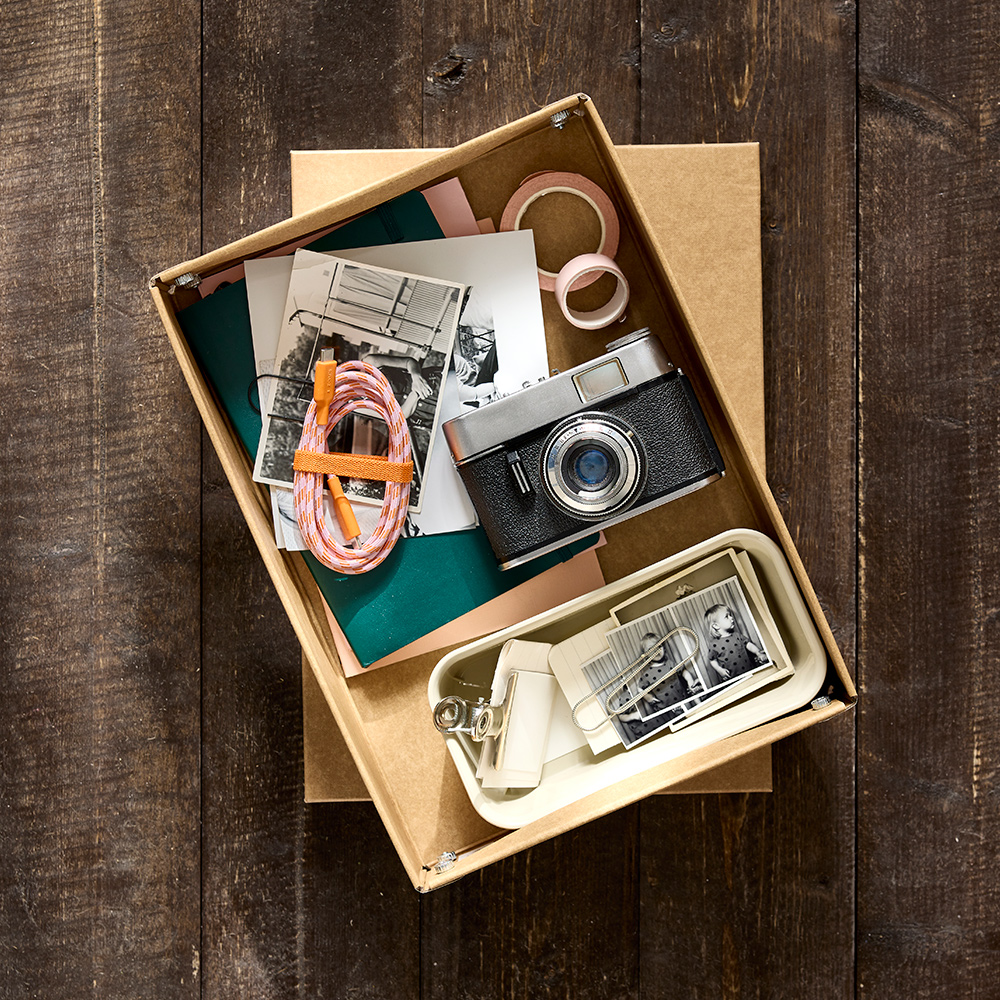 Una caja de almacenamiento de papel GÄRDESGÅRD, con la tapa retirada, colocada sobre un suelo de madera y que contiene accesorios y materiales de fotografía.