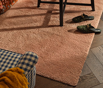 En el suelo del salón hay una alfombra STOENSE de color rosa pálido. Sobre ella hay una mesa de centro negra y, junto a ella, un sillón a cuadros.