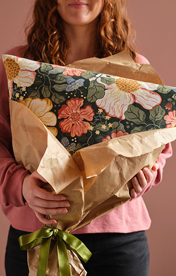 Una persona en una habitación rosa sostiene un cojín con una funda floral de ARENBOK envuelta en papel marrón y atada con una cinta verde.