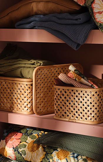 Las cajas VIELÅNGEN de color beige, colocadas en un soporte de bambú, se encuentran dentro de un armario rosa junto con ropa de cama de colores.